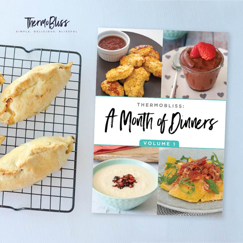 A ThermoBliss Month of Dinners cookbook next to bread rolls on a baking tray.